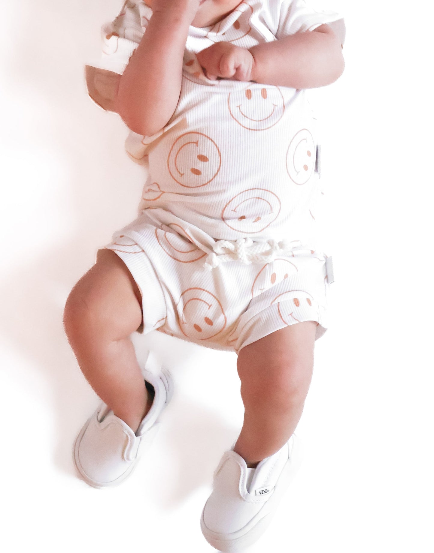 Photo of a baby boy wearing a white shirt and shorties set adorned with camel-colored smiley faces, ideal for toddlers. The design features ribbed bamboo fabric, ensuring comfort and breathability. Available sizes include 3-6 months, 6-12 months, 12-18 months, and 18-24 months, making it a versatile and stylish choice for both boys and girls.