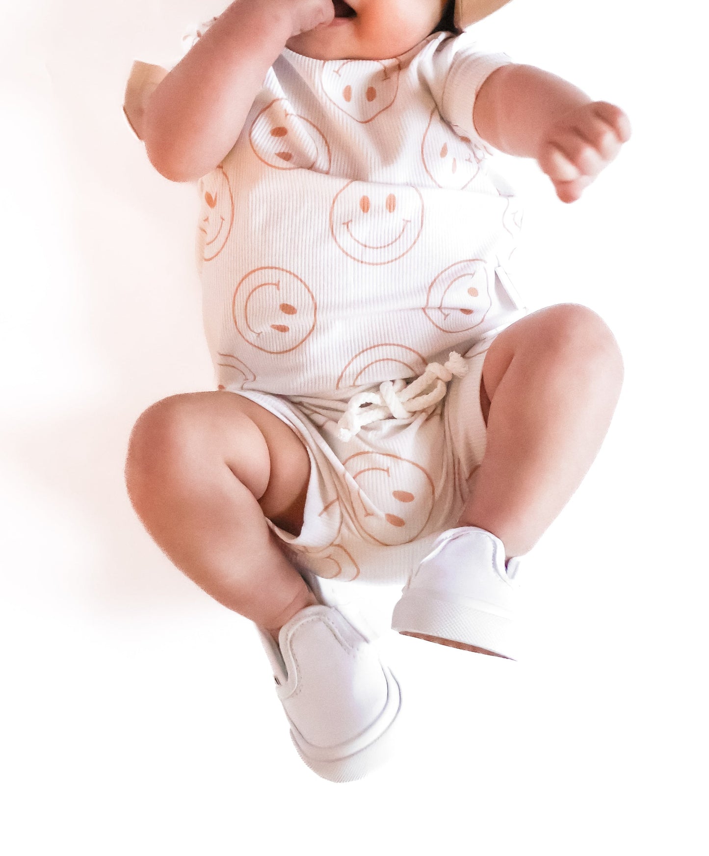 Photo of a baby boy wearing a white shirt and shorties set adorned with camel-colored smiley faces, ideal for toddlers. The design features ribbed bamboo fabric, ensuring comfort and breathability. Available sizes include 3-6 months, 6-12 months, 12-18 months, and 18-24 months, making it a versatile and stylish choice for both boys and girls.