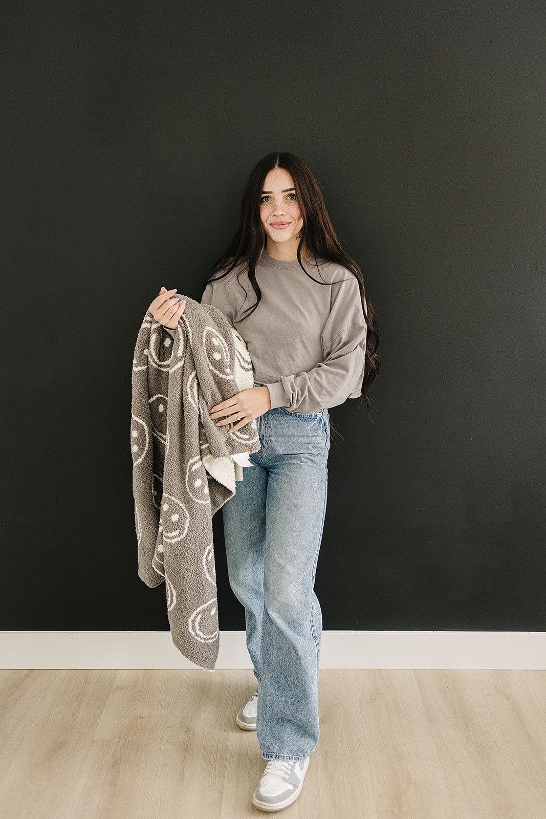 Photo of a woman holding a very cozy charcoal smiley plush blanket measuring 45x60 inches. The blanket features a charcoal grey background with white smiley faces on one side, and the reverse design with a white background and charcoal grey smiley faces on the other side. This soft and inviting blanket adds a touch of fun and warmth to any space.