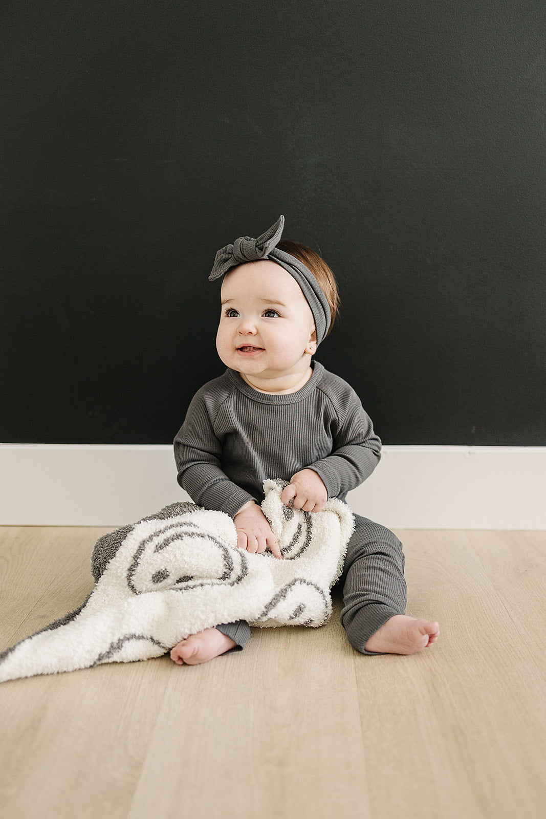 Photo of a baby holding a very cozy charcoal smiley plush blanket measuring 45x60 inches. The blanket features a charcoal grey background with white smiley faces on one side, and the reverse design with a white background and charcoal grey smiley faces on the other side. This soft and inviting blanket adds a touch of fun and warmth to any space.