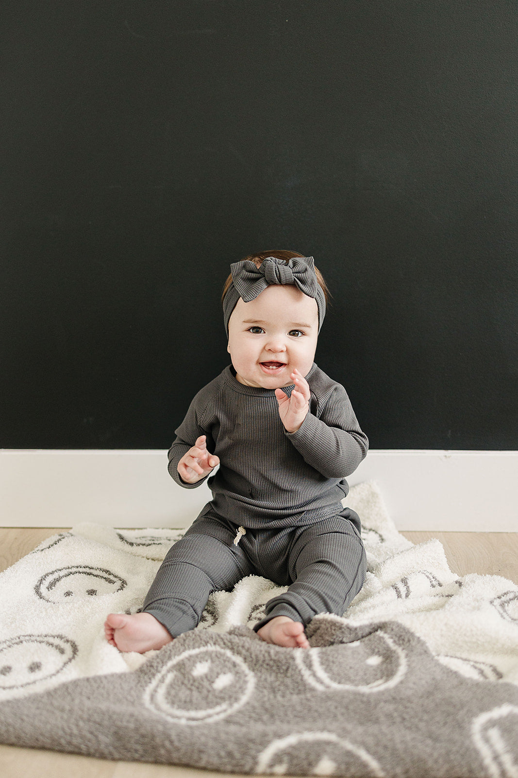 Photo of a baby girl sitting on a very cozy charcoal smiley plush blanket measuring 45x60 inches. The blanket features a charcoal grey background with white smiley faces on one side, and the reverse design with a white background and charcoal grey smiley faces on the other side. This soft and inviting blanket adds a touch of fun and warmth to any space.