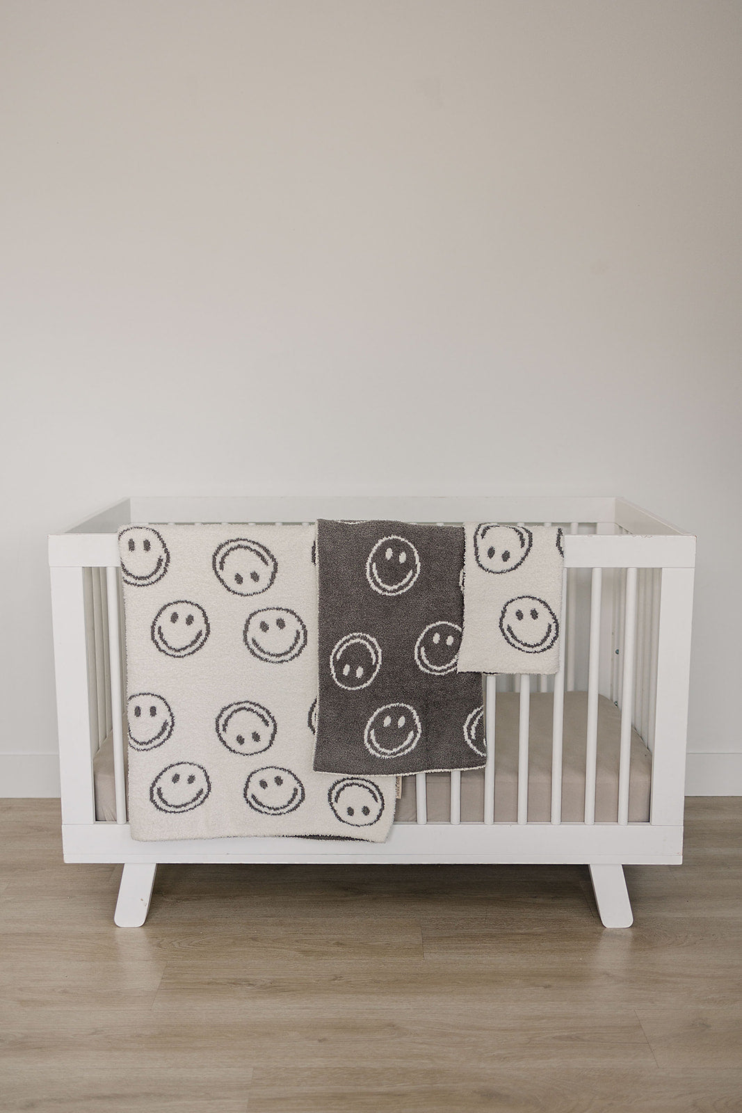 Photo of a white crib holding a very cozy charcoal smiley plush blanket measuring 45x60 inches. The blanket features a charcoal grey background with white smiley faces on one side, and the reverse design with a white background and charcoal grey smiley faces on the other side. This soft and inviting blanket adds a touch of fun and warmth to any space.