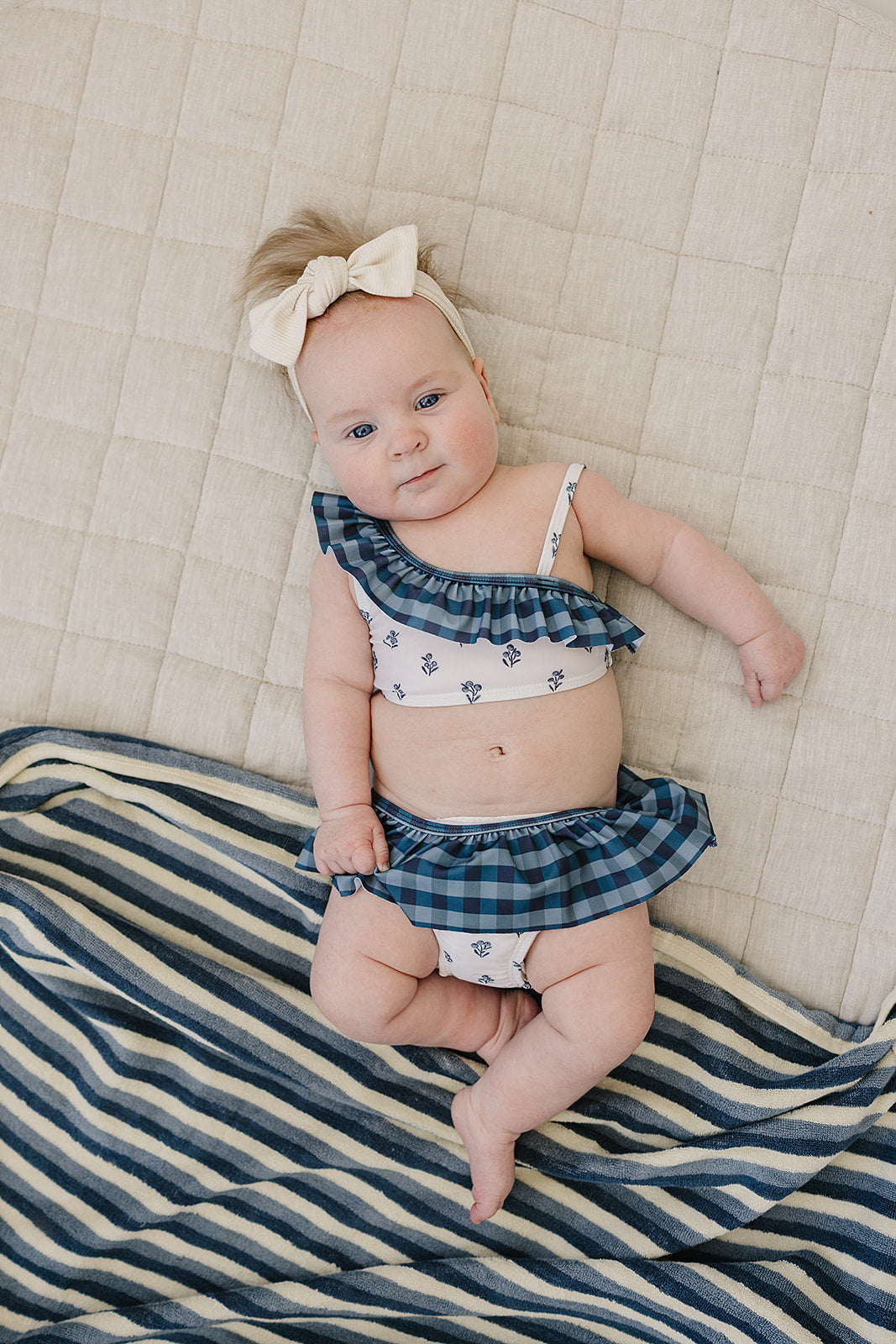 Baby wearing the Mebie Baby Cream Berry Ruffle Shoulder Bikini Set, a blue plaid and floral two-piece with ruffle accents, lying on a soft beige blanket with a striped towel nearby.