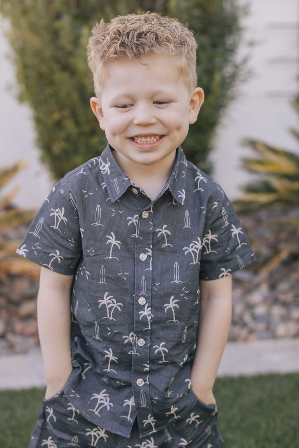 Young boy wearing the Mebie Baby Navy Surfer Linen Button Up Shirt, a dark blue short-sleeve shirt with palm tree and surfboard print, smiling with hands in pockets.