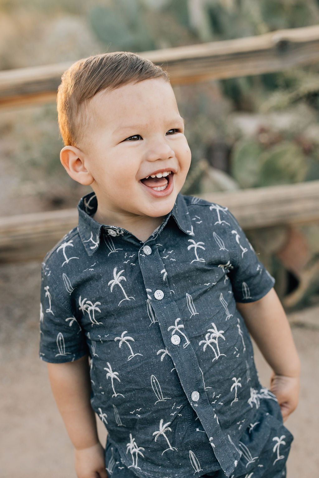Young boy laughing and wearing the Mebie Baby Navy Surfer Linen Button Up Shirt, featuring a navy background with white palm tree and surfboard pattern.