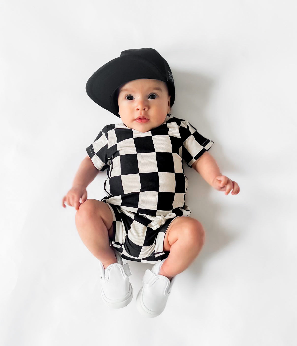 Photo of a baby boy wearing a stylish black and white checkered shirt and shorties set for toddlers. The design features a ribbed bamboo fabric, offering comfort and breathability. Available in sizes 3-6 months, 6-12 months, 12-18 months, and 18-24 months, this outfit is perfect for both boys and girls, combining trendy patterns with eco-friendly materials.