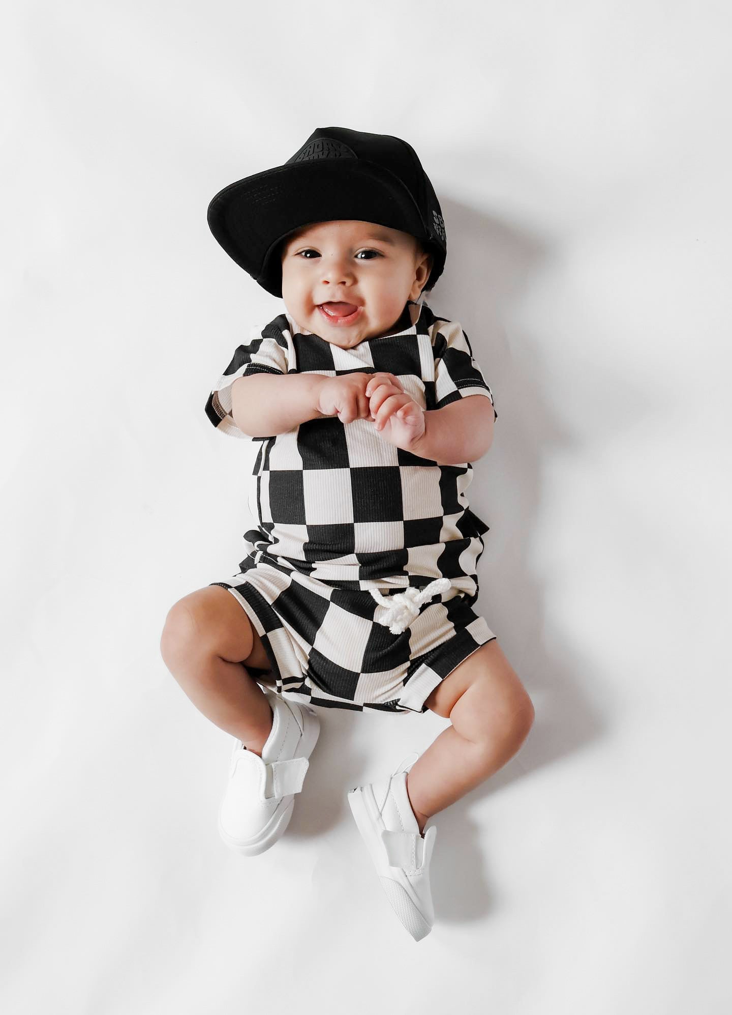 Photo of a baby boy wearing a stylish black and white checkered shirt and shorties set for toddlers. The design features a ribbed bamboo fabric, offering comfort and breathability. Available in sizes 3-6 months, 6-12 months, 12-18 months, and 18-24 months, this outfit is perfect for both boys and girls, combining trendy patterns with eco-friendly materials.