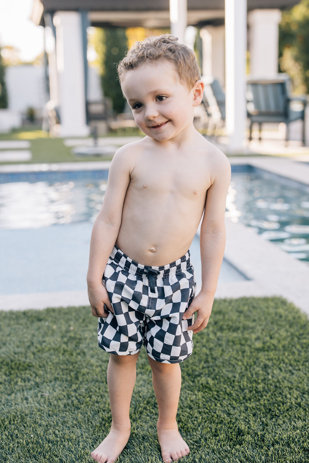 Young boy wearing Mebie Baby Wavy Checkered Swim Shorts, black and white checkered swim trunks, smiling by the poolside on green grass.