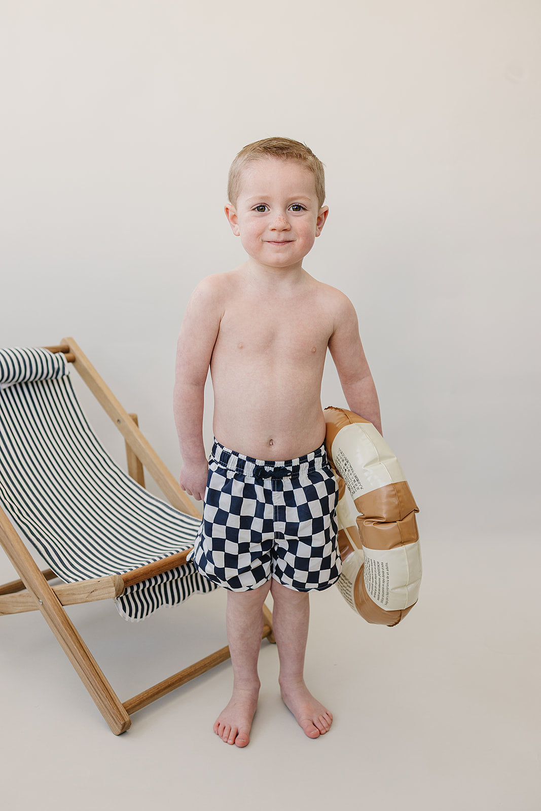 Young boy wearing Mebie Baby Wavy Checkered Swim Shorts, holding a pool float next to a striped beach chair in a studio setting.