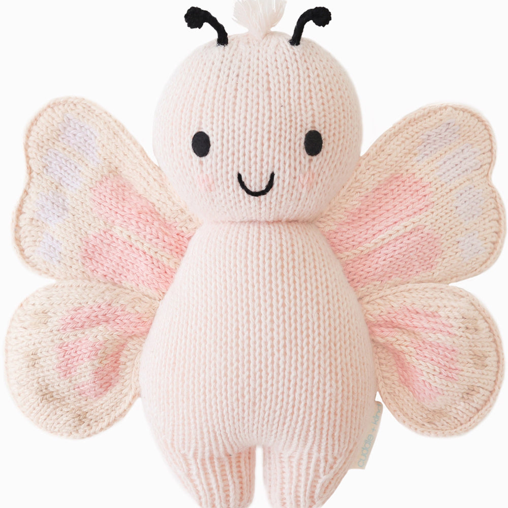 Cuddle + Kind Baby Butterfly (Petal Pink), hand-knit plush butterfly with smiling face and pastel pink wings, shown on a white background.