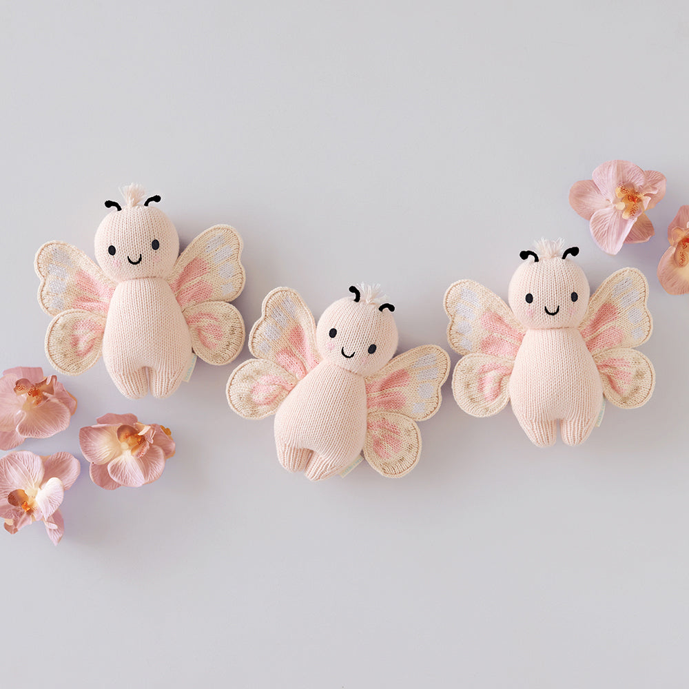 
                  
                    Three Cuddle + Kind Baby Butterfly (Petal Pink) plush toys arranged with pink flowers on a light background.
                  
                