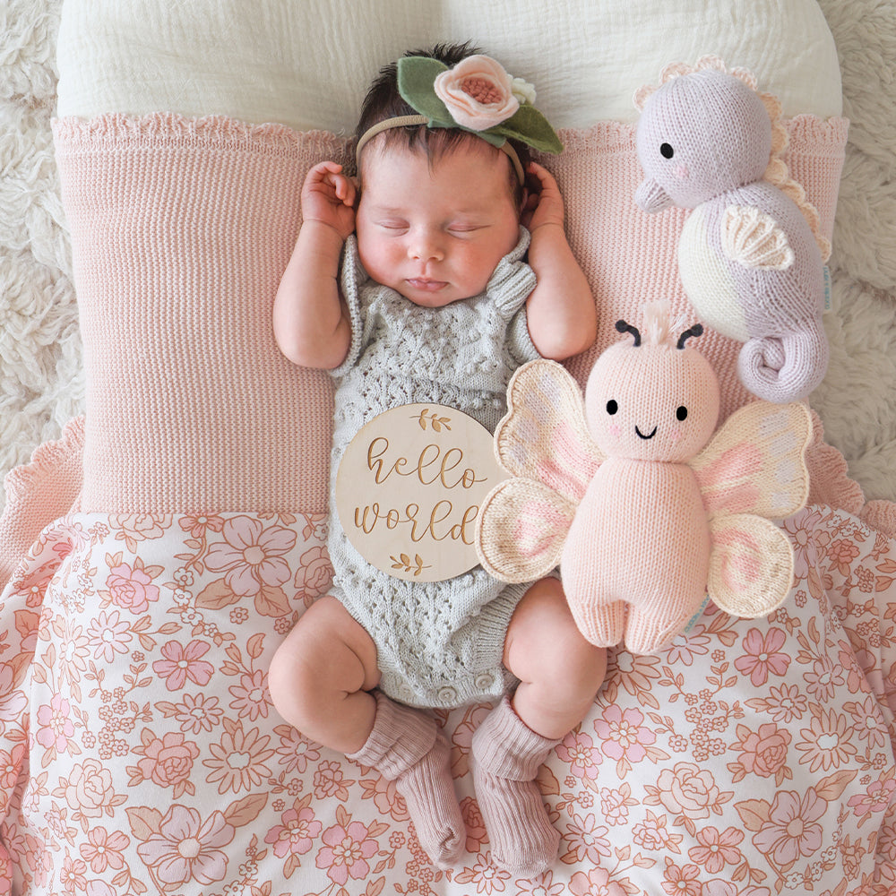
                  
                    Baby sleeping on floral bedding with Cuddle + Kind Baby Butterfly (Petal Pink) and Baby Seahorse (Lilac) plush toys and a wooden “Hello World” sign.
                  
                