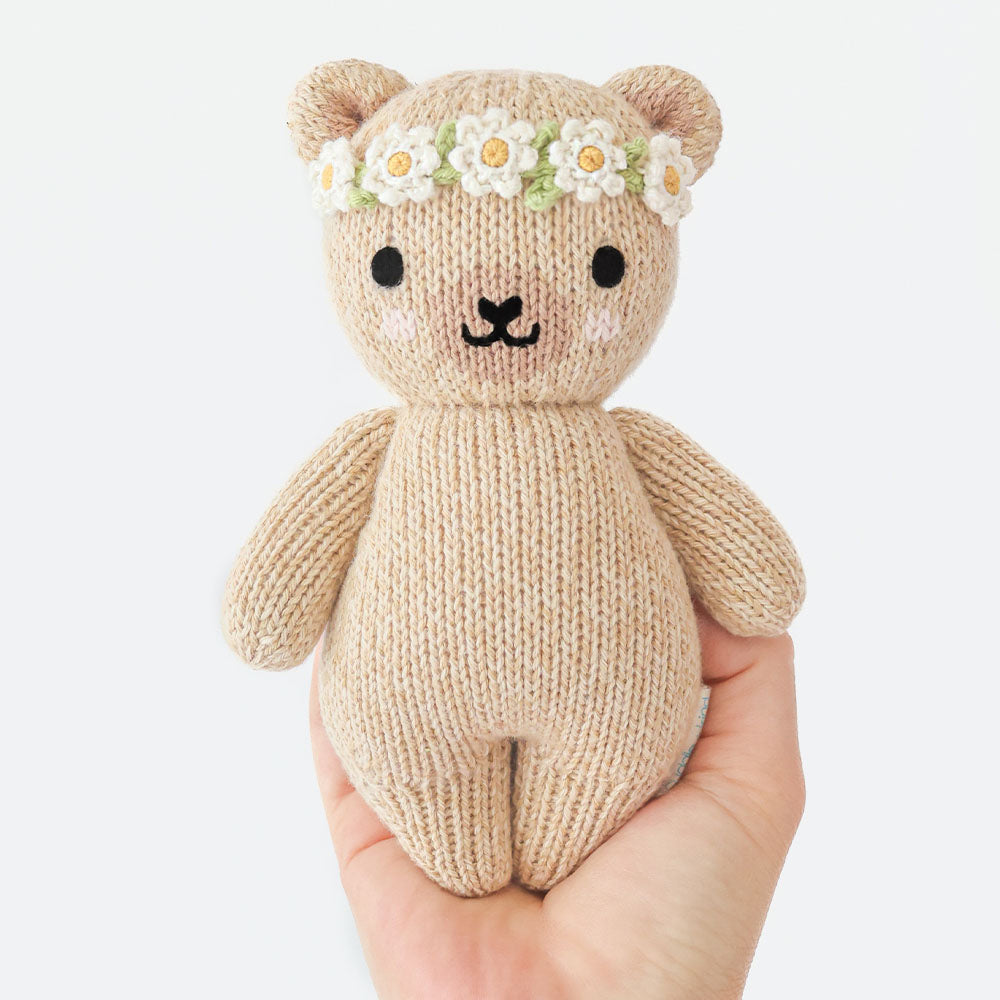 
                  
                    Hand holding the Cuddle + Kind Baby Honey Bear (Ivory Floral), a small hand-knit teddy bear with ivory floral crown and embroidered face.
                  
                