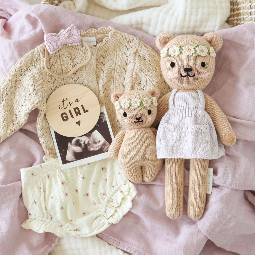 
                  
                    Styled flat lay featuring Cuddle + Kind Baby Honey Bear (Ivory Floral) and matching larger bear, surrounded by baby clothes, a wooden “It’s a Girl” sign, and a pink blanket.
                  
                