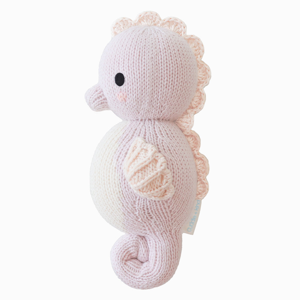 Side view of Cuddle + Kind Baby Seahorse (Lilac), hand-knit lilac seahorse doll with cream belly and pink details, displayed on a white background.