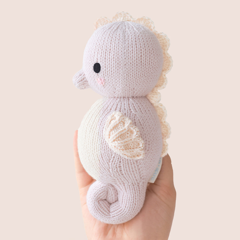 
                  
                    Side view of Cuddle + Kind Baby Seahorse (Lilac), hand-knit seahorse plush with curled tail and textured back, shown being held in a hand.
                  
                