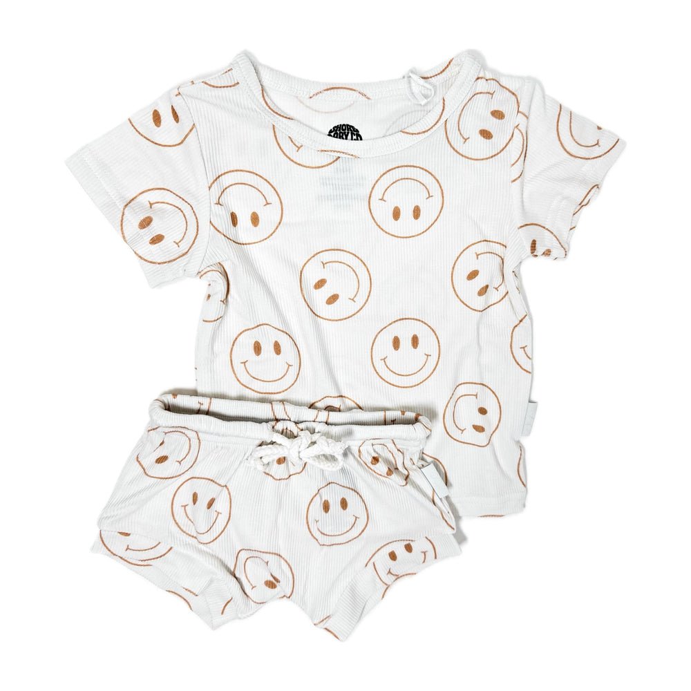 Photo of a white shirt and shorties set adorned with camel-colored smiley faces, ideal for toddlers. The design features ribbed bamboo fabric, ensuring comfort and breathability. Available sizes include 3-6 months, 6-12 months, 12-18 months, and 18-24 months, making it a versatile and stylish choice for both boys and girls.