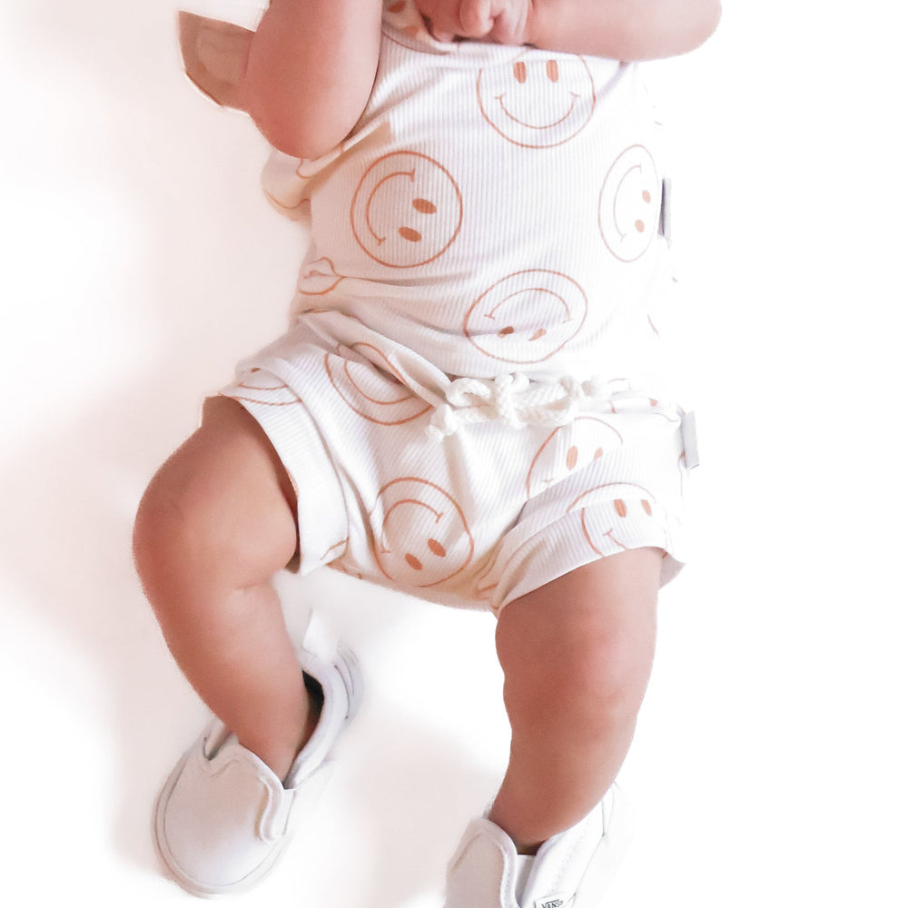 
                  
                    Photo of a baby boy wearing a white shirt and shorties set adorned with camel-colored smiley faces, ideal for toddlers. The design features ribbed bamboo fabric, ensuring comfort and breathability. Available sizes include 3-6 months, 6-12 months, 12-18 months, and 18-24 months, making it a versatile and stylish choice for both boys and girls.
                  
                