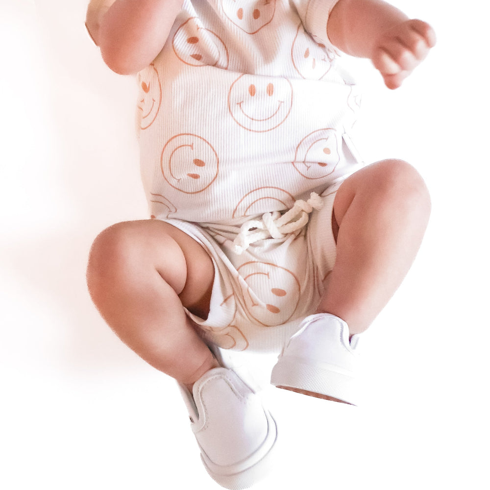 
                  
                    Photo of a baby boy wearing a white shirt and shorties set adorned with camel-colored smiley faces, ideal for toddlers. The design features ribbed bamboo fabric, ensuring comfort and breathability. Available sizes include 3-6 months, 6-12 months, 12-18 months, and 18-24 months, making it a versatile and stylish choice for both boys and girls.
                  
                