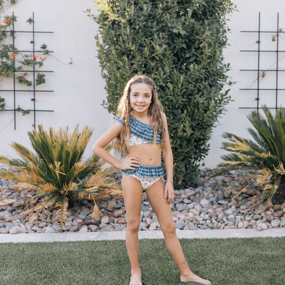 
                  
                    Young girl wearing the Mebie Baby Cream Berry Ruffle Shoulder Bikini Set, a blue plaid and floral two-piece swimsuit with one-shoulder ruffle top, standing confidently in a backyard garden.
                  
                