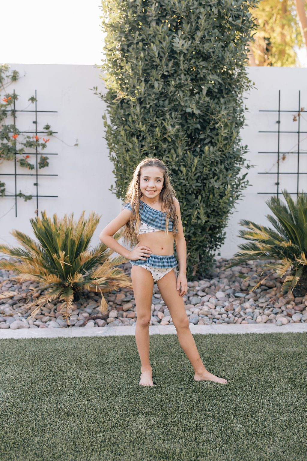 
                  
                    Young girl wearing the Mebie Baby Cream Berry Ruffle Shoulder Bikini Set, a blue plaid and floral two-piece swimsuit with one-shoulder ruffle top, standing confidently in a backyard garden.
                  
                
