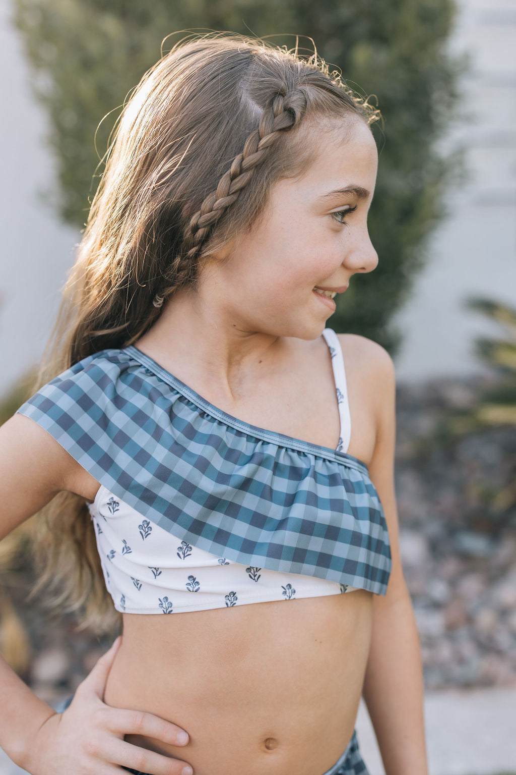 
                  
                    Close-up of girl in the Mebie Baby Cream Berry Ruffle Shoulder Bikini Set, showing blue plaid and floral print, ruffle detail, and braided hairstyle.
                  
                