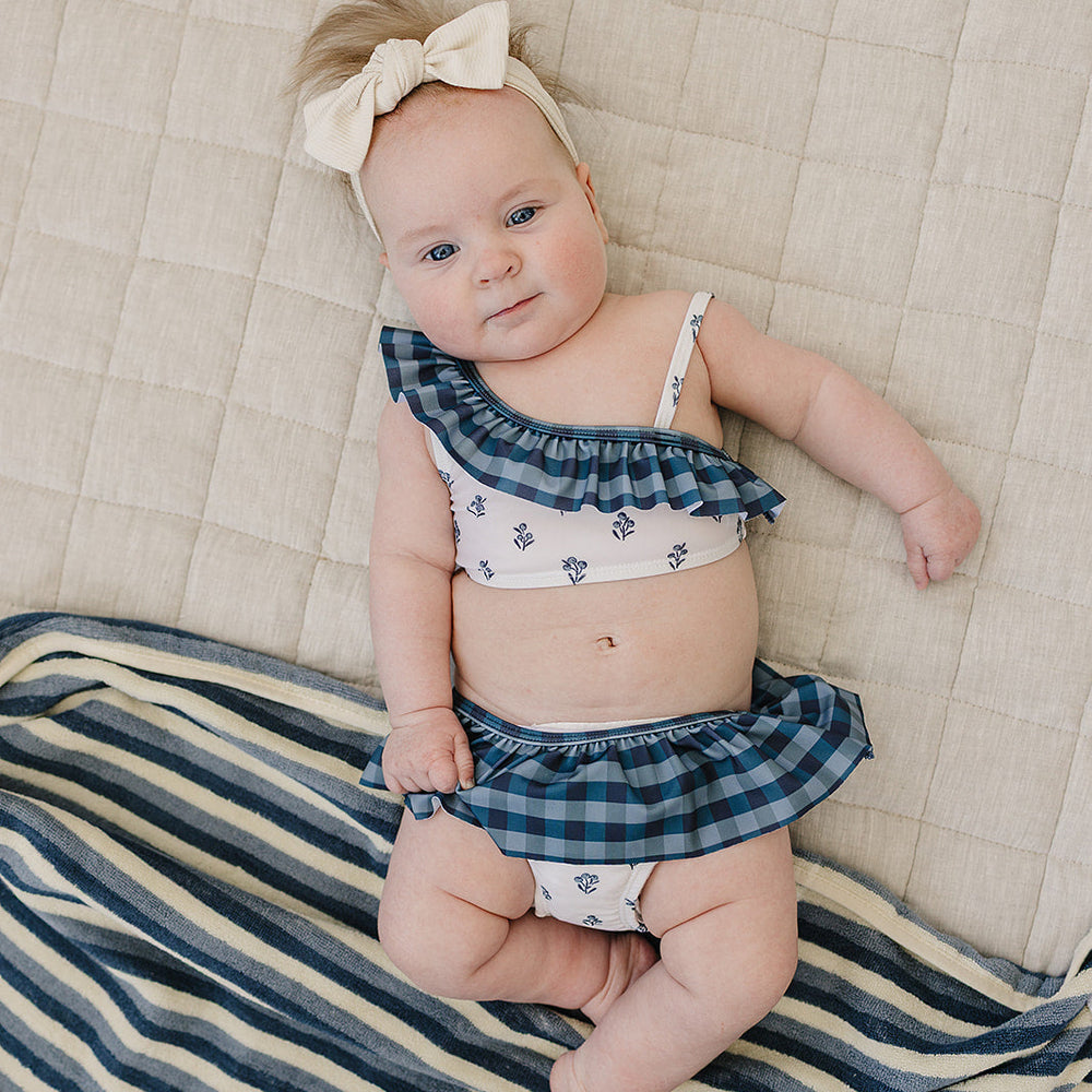 
                  
                    Baby wearing the Mebie Baby Cream Berry Ruffle Shoulder Bikini Set, a blue plaid and floral two-piece with ruffle accents, lying on a soft beige blanket with a striped towel nearby.
                  
                