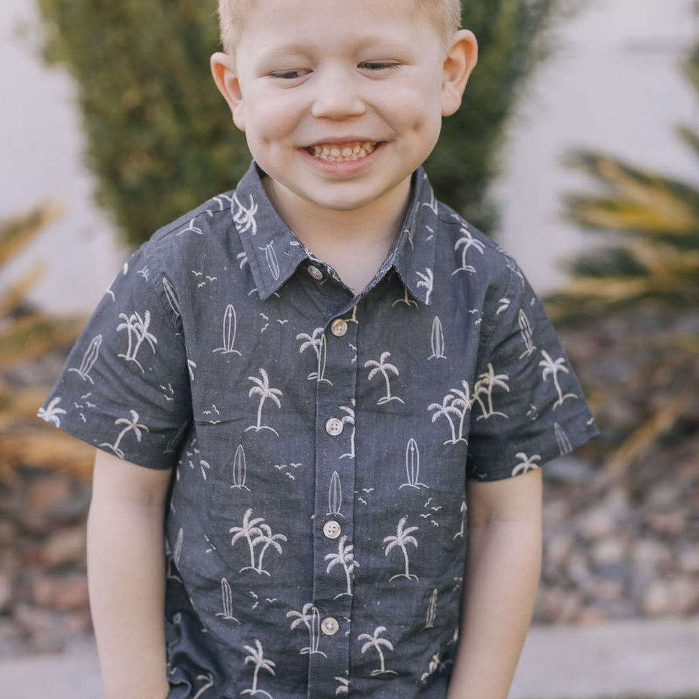
                  
                    Young boy wearing the Mebie Baby Navy Surfer Linen Button Up Shirt, a dark blue short-sleeve shirt with palm tree and surfboard print, smiling with hands in pockets.
                  
                