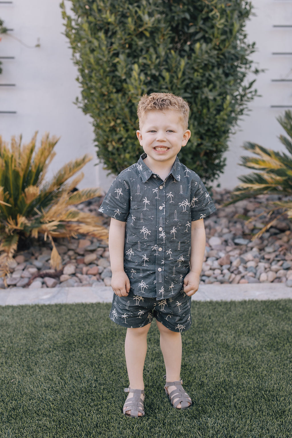 
                  
                    Young boy standing outdoors wearing the Mebie Baby Navy Surfer Linen Button Up Shirt and matching shorts, smiling on green grass with plants in the background.
                  
                