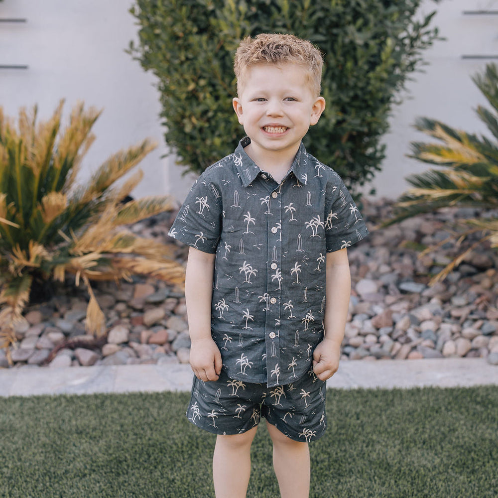 
                  
                    Young boy wearing the Mebie Baby Navy Surfer Linen Cotton Shorts paired with the matching button-up shirt, standing on green grass and smiling outdoors.
                  
                