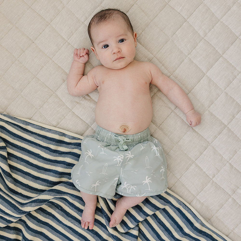 
                  
                    Baby wearing Mebie Baby Sage Surfer Swim Shorts, light green swim trunks with palm tree and surfboard print, lying on a soft beige quilt with a striped towel.
                  
                