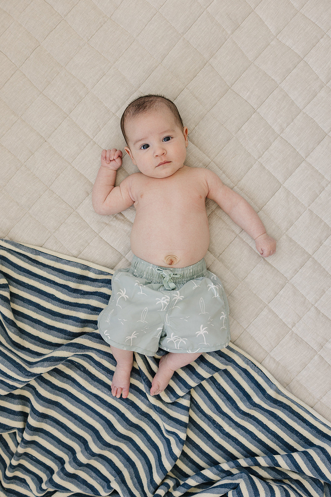 
                  
                    Baby wearing Mebie Baby Sage Surfer Swim Shorts, light green swim trunks with palm tree and surfboard print, lying on a soft beige quilt with a striped towel.
                  
                