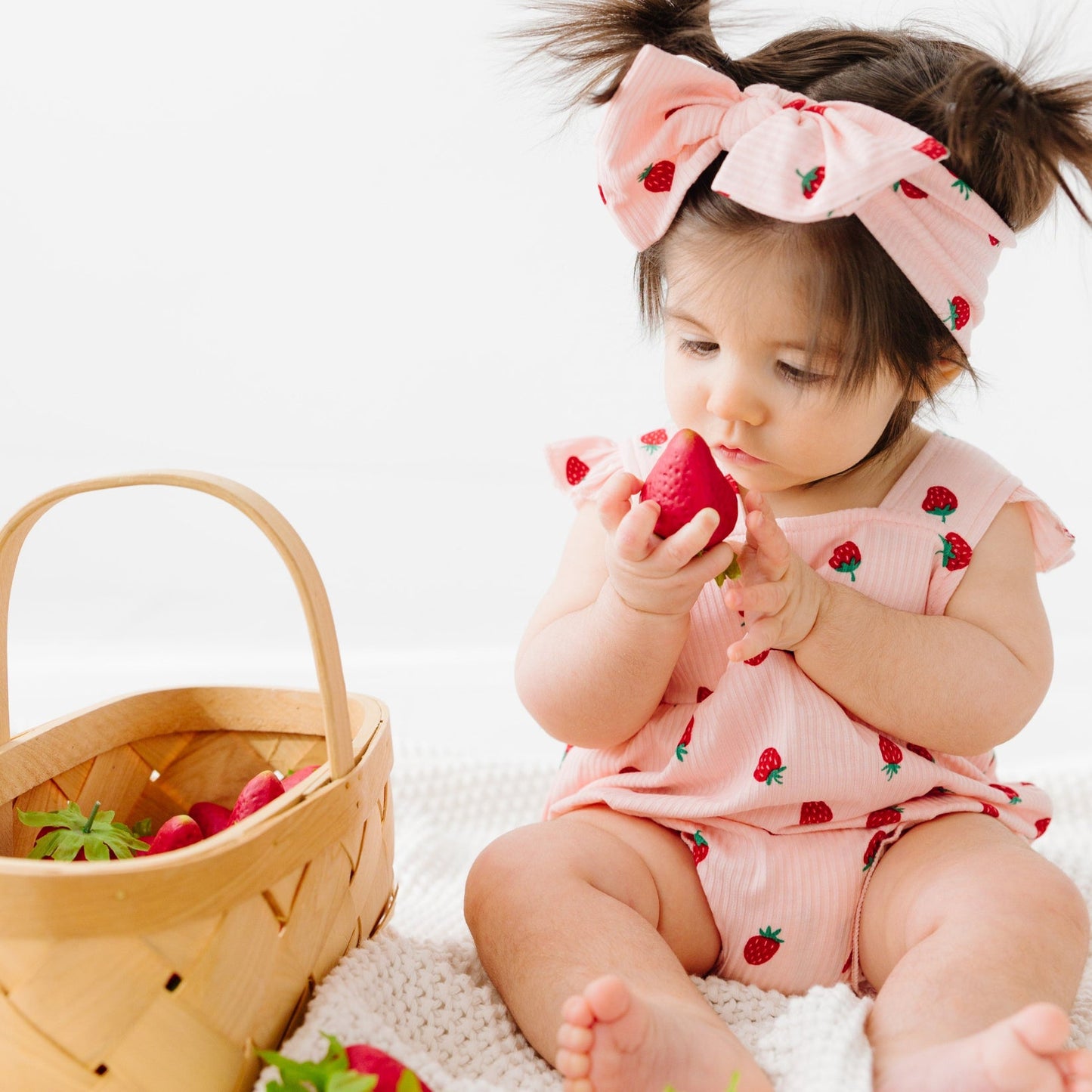 
                  
                    Baby girl wearing pink strawberry romper and matching bow headband, holding a strawberry next to a wooden basket.
                  
                