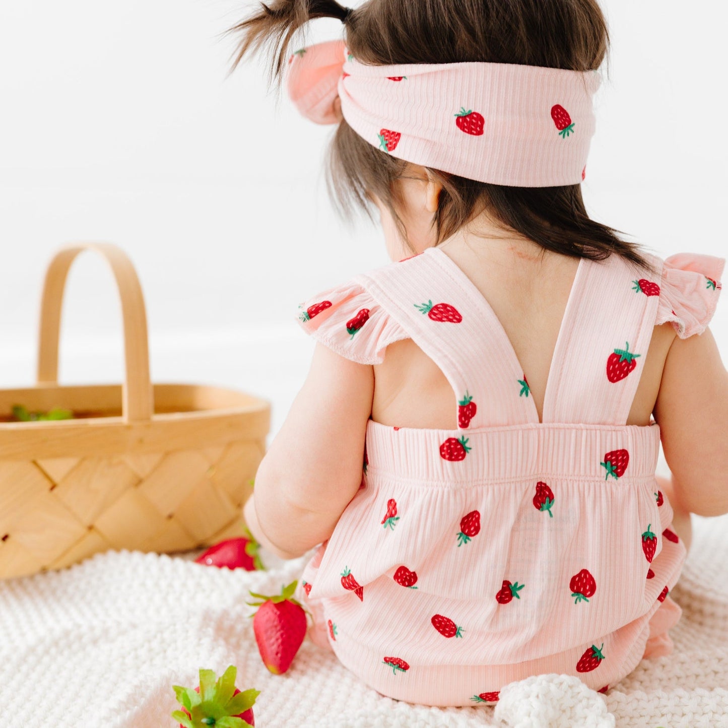 
                  
                    Back view of toddler girl in pink strawberry romper with criss-cross straps and matching bow headband, sitting by a basket.
                  
                
