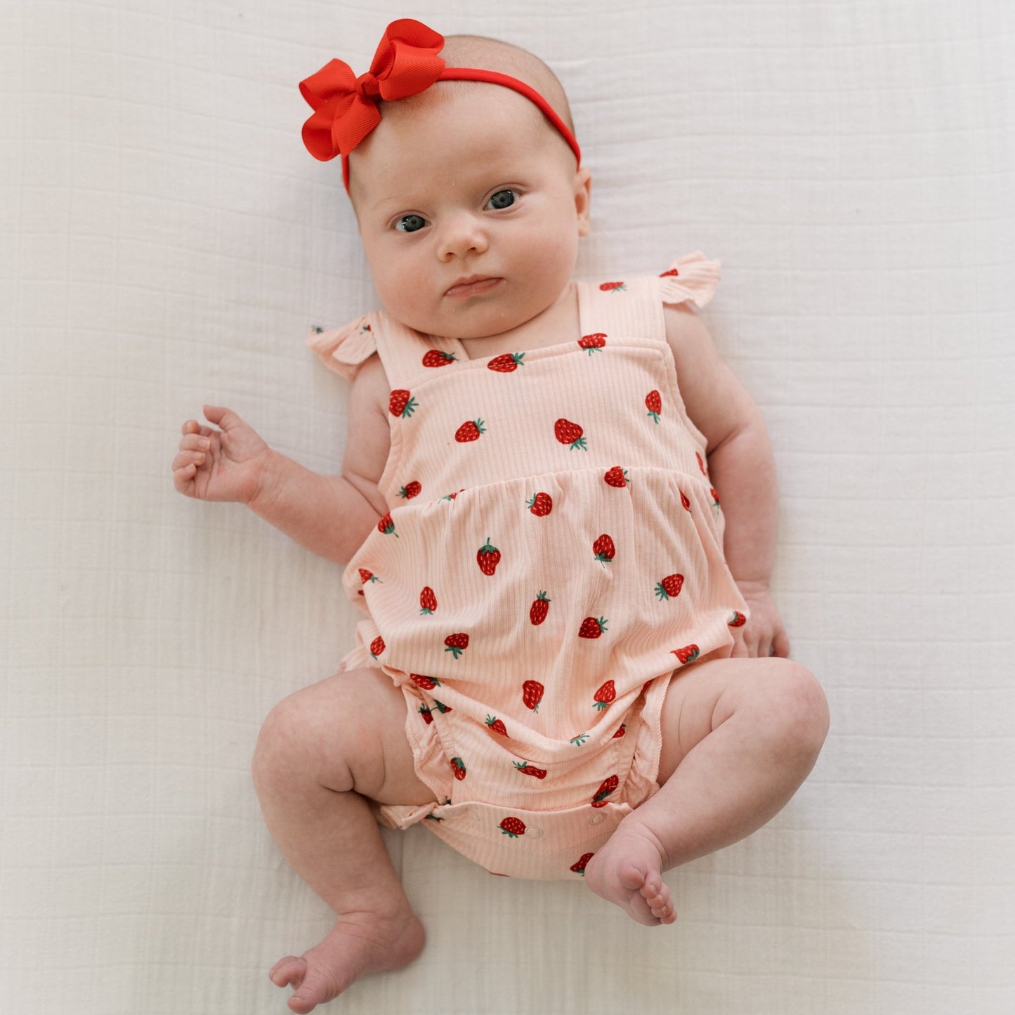 
                  
                    Baby girl wearing strawberry print romper with flutter sleeves and red bow headband, lying on white blanket.
                  
                