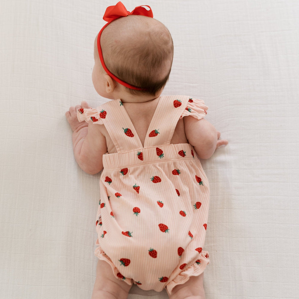 
                  
                    Back of baby girl wearing pink romper with red strawberries and criss-cross straps, paired with red bow headband.
                  
                