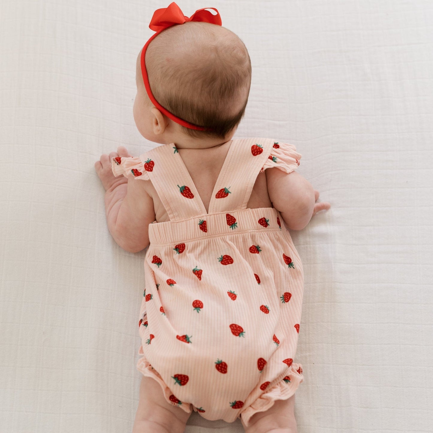 
                  
                    Back of baby girl wearing pink romper with red strawberries and criss-cross straps, paired with red bow headband.
                  
                