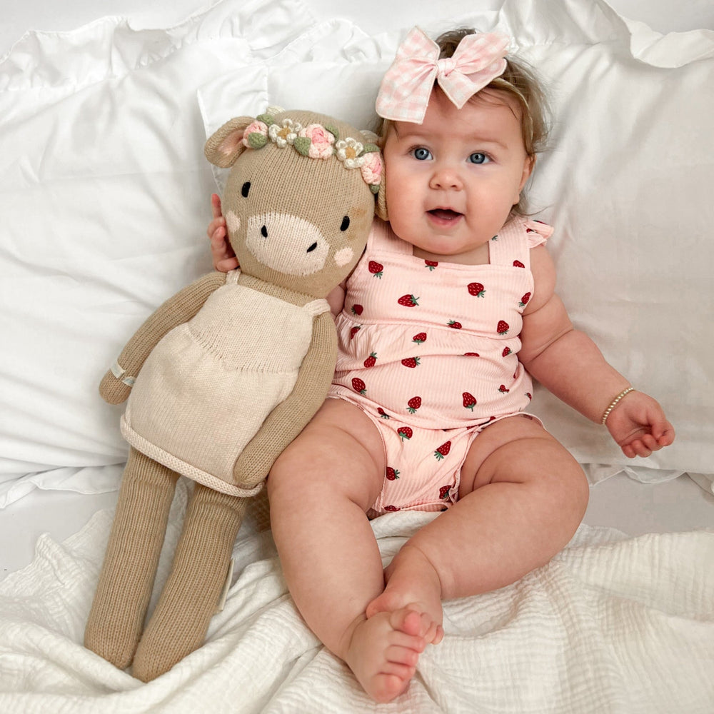 
                  
                    Smiling baby girl in strawberry romper with pink plaid bow, sitting next to a knit floral crown doll on white bedding.
                  
                