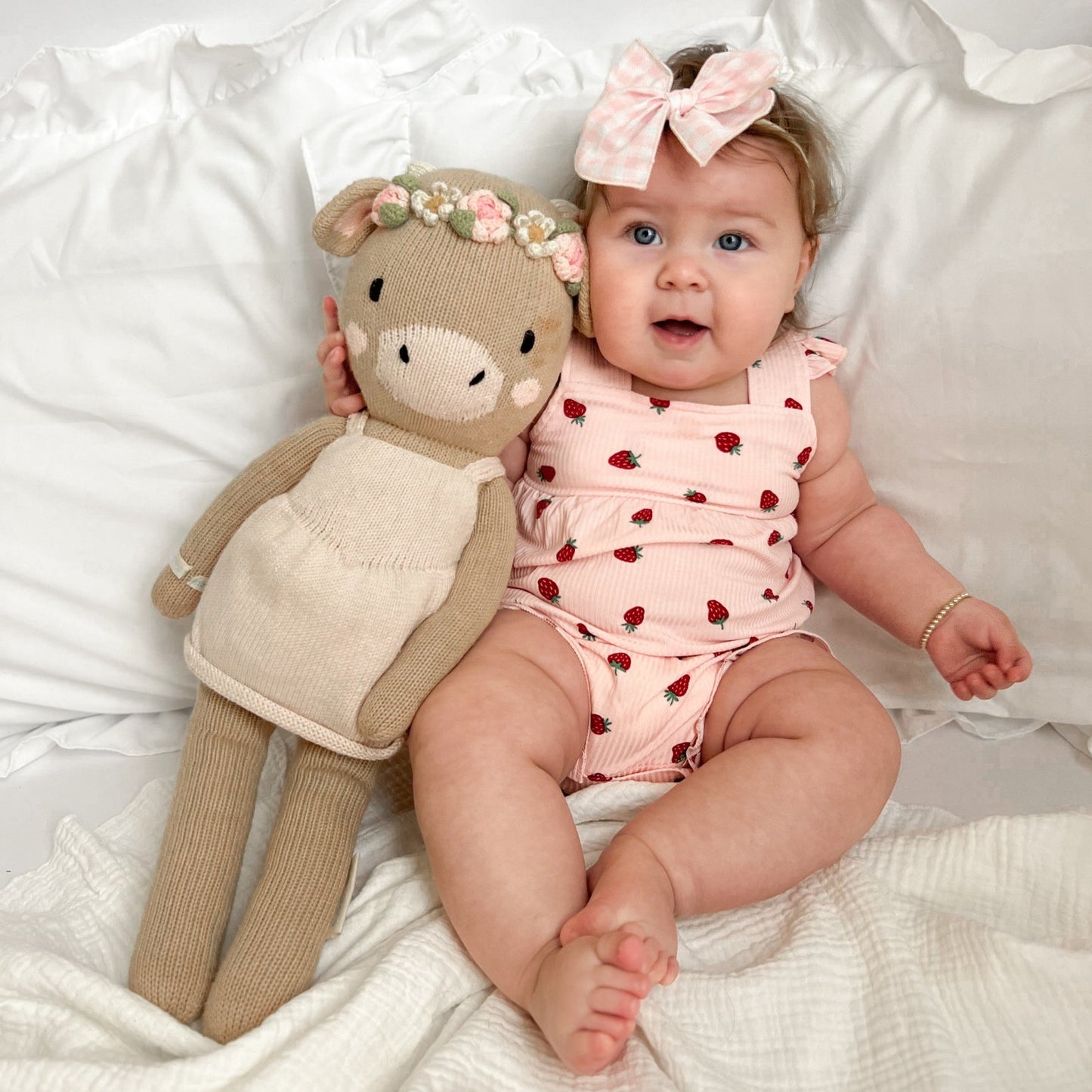 
                  
                    Smiling baby girl in strawberry romper with pink plaid bow, sitting next to a knit floral crown doll on white bedding.
                  
                
