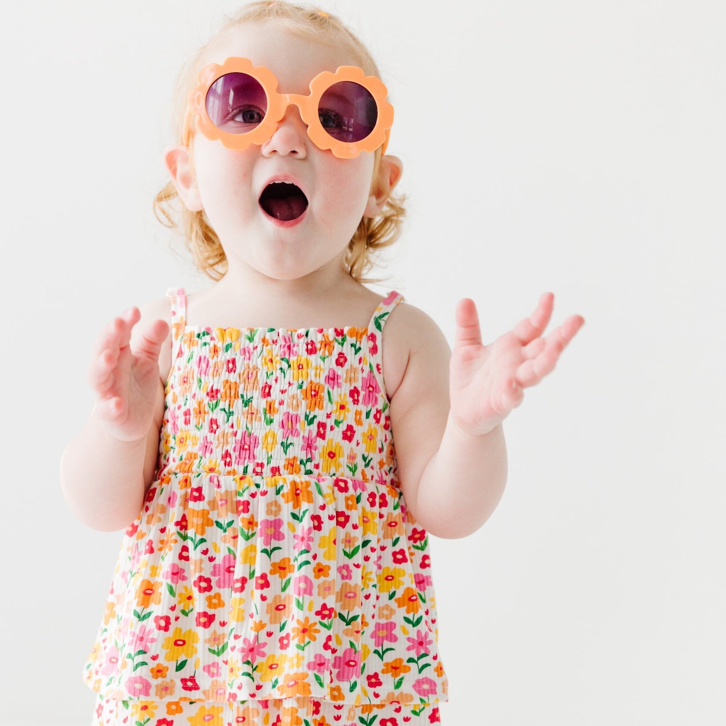 
                  
                    Little girl wearing smocked floral romper with spaghetti straps and orange flower-shaped sunglasses, smiling in excitement.
                  
                