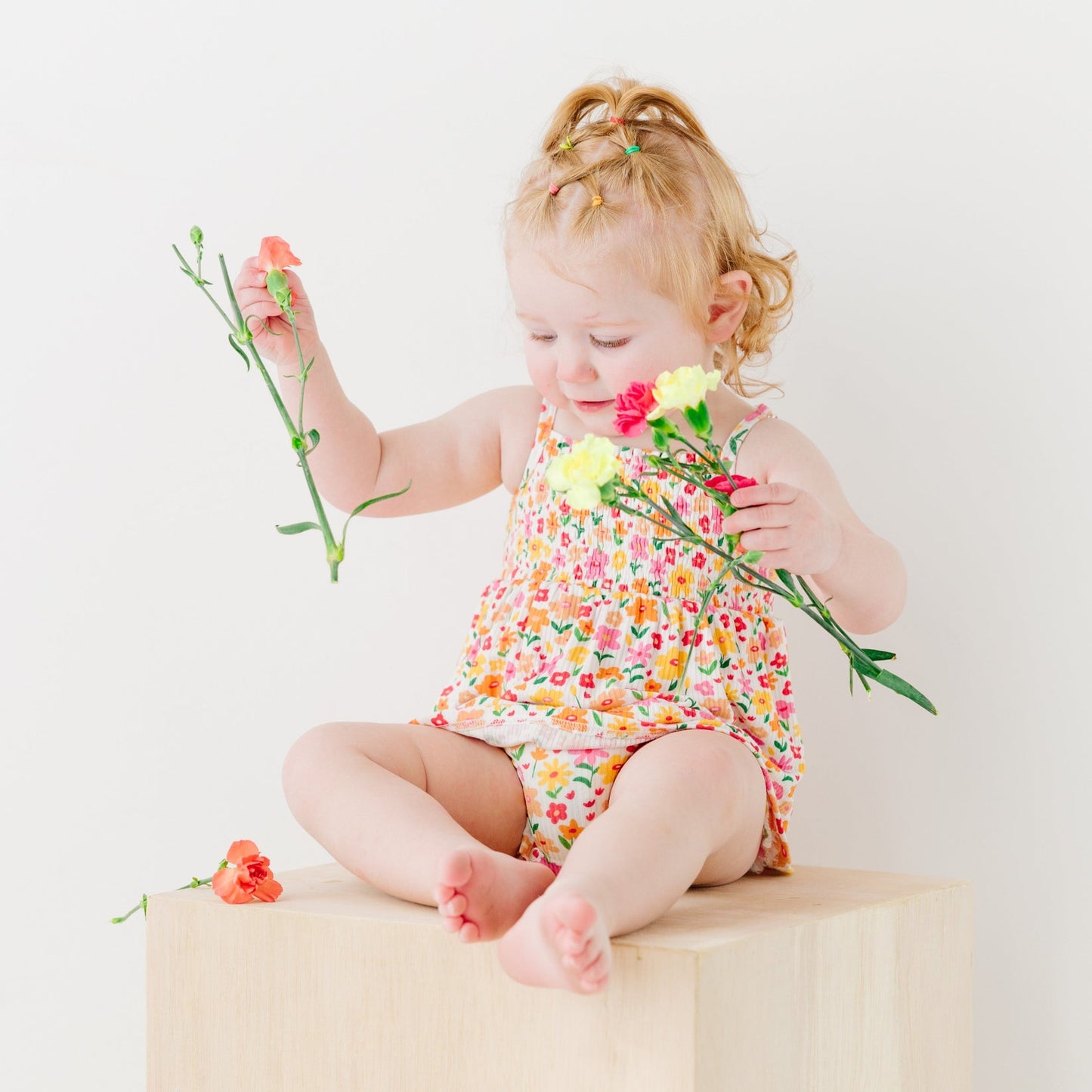 
                  
                    Toddler girl wearing Summer Blooms floral romper, holding yellow and orange flowers while seated on a light wood cube.
                  
                