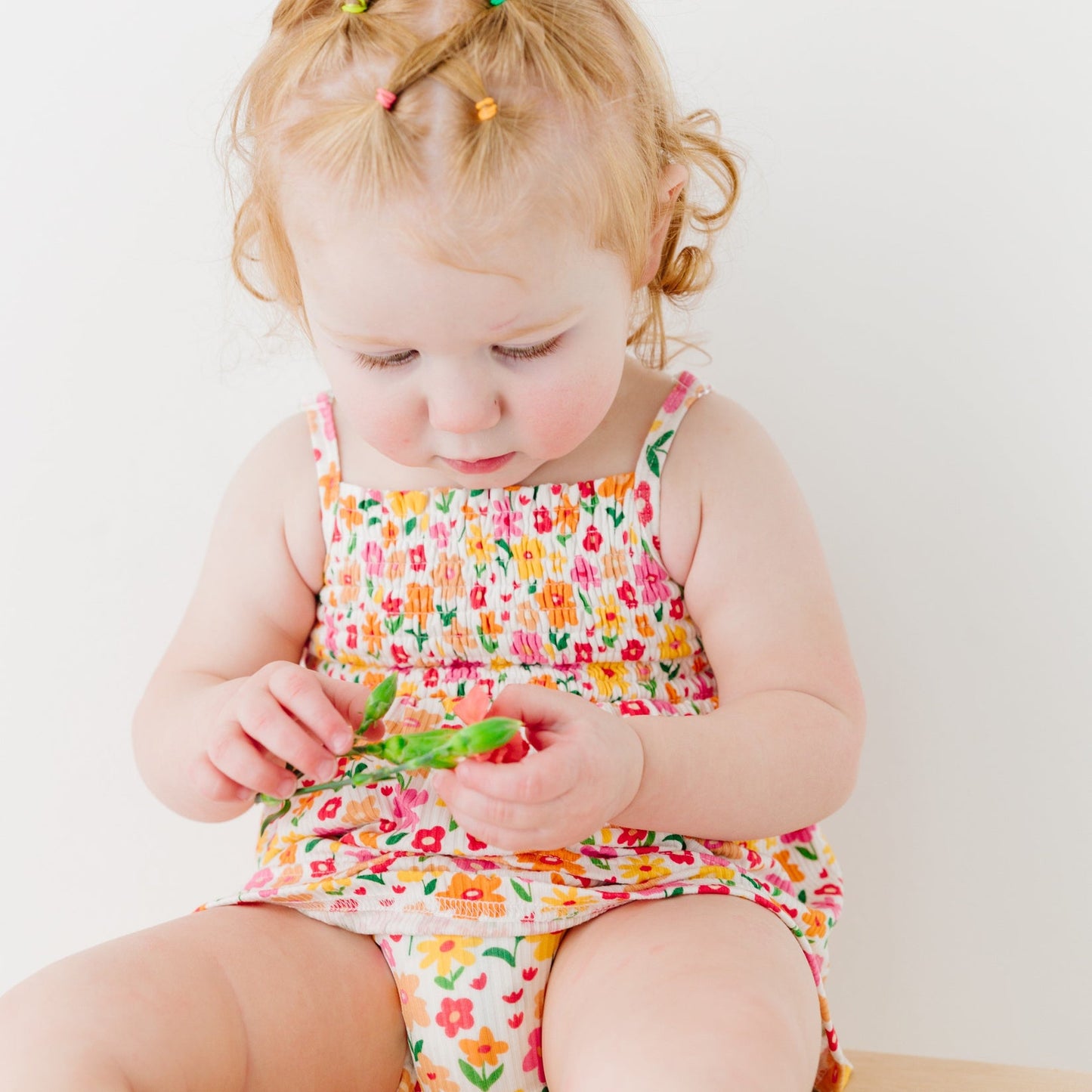 
                  
                    Close-up of toddler in colorful floral romper holding a small flower, with detailed view of smocked bodice.
                  
                