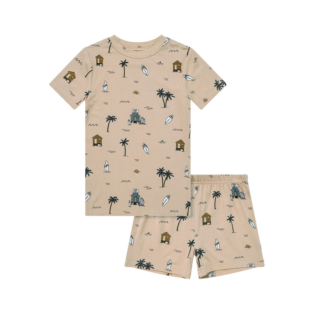 Flat lay of Surf Shack two-piece set for toddlers – tan shirt and shorts printed with palm trees, surfboards, waves, and beach huts.
