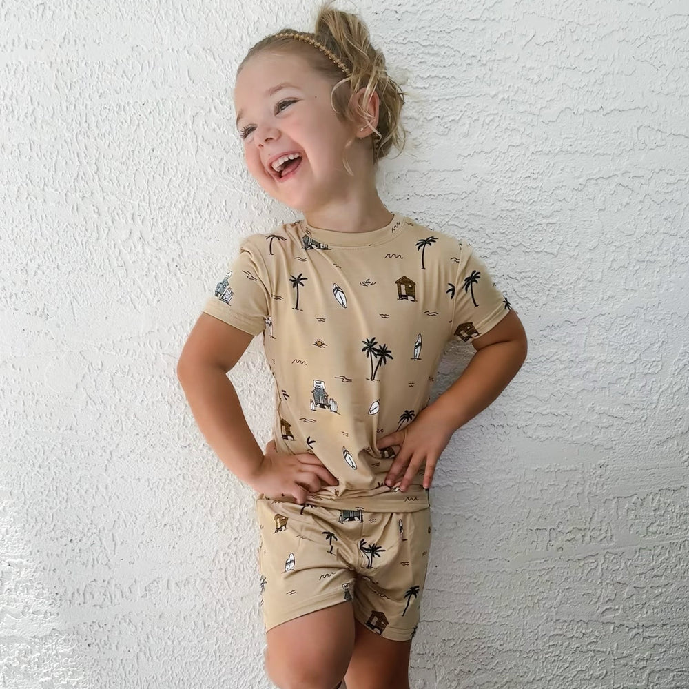 
                  
                    Laughing toddler girl wearing tan Surf Shack two-piece outfit with surfboards, huts, and palm tree illustrations.
                  
                