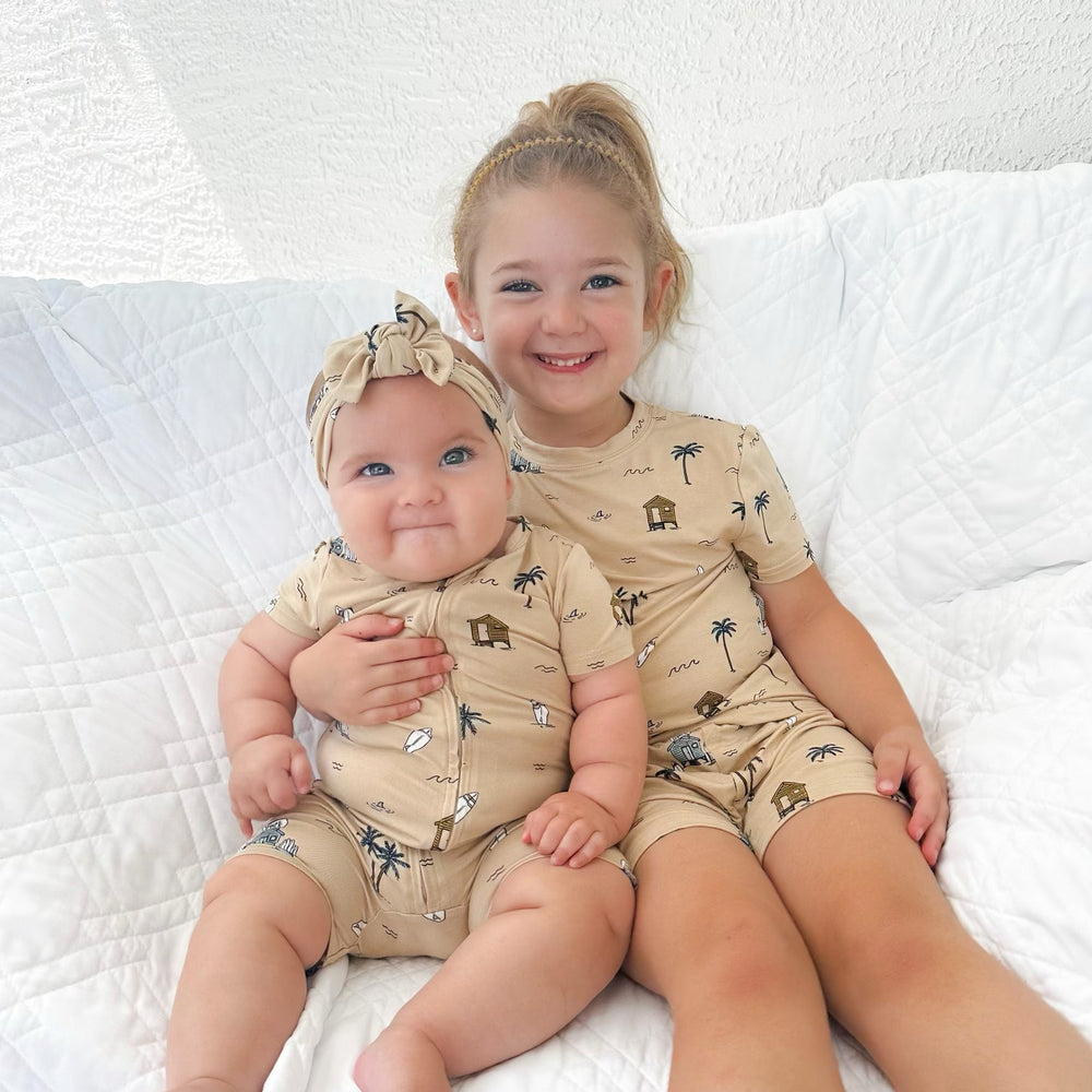 
                  
                    Big sister and baby girl wearing matching Surf Shack summer outfits in tan, featuring beach huts, surfboards, and palm trees – sibling photo.
                  
                