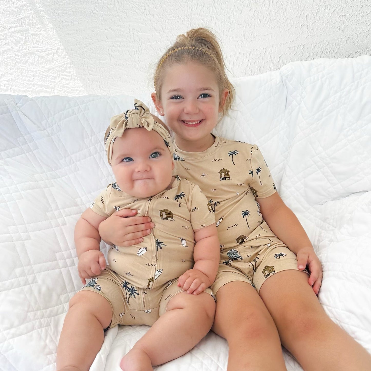 
                  
                    Big sister and baby girl wearing matching Surf Shack summer outfits in tan, featuring beach huts, surfboards, and palm trees – sibling photo.
                  
                