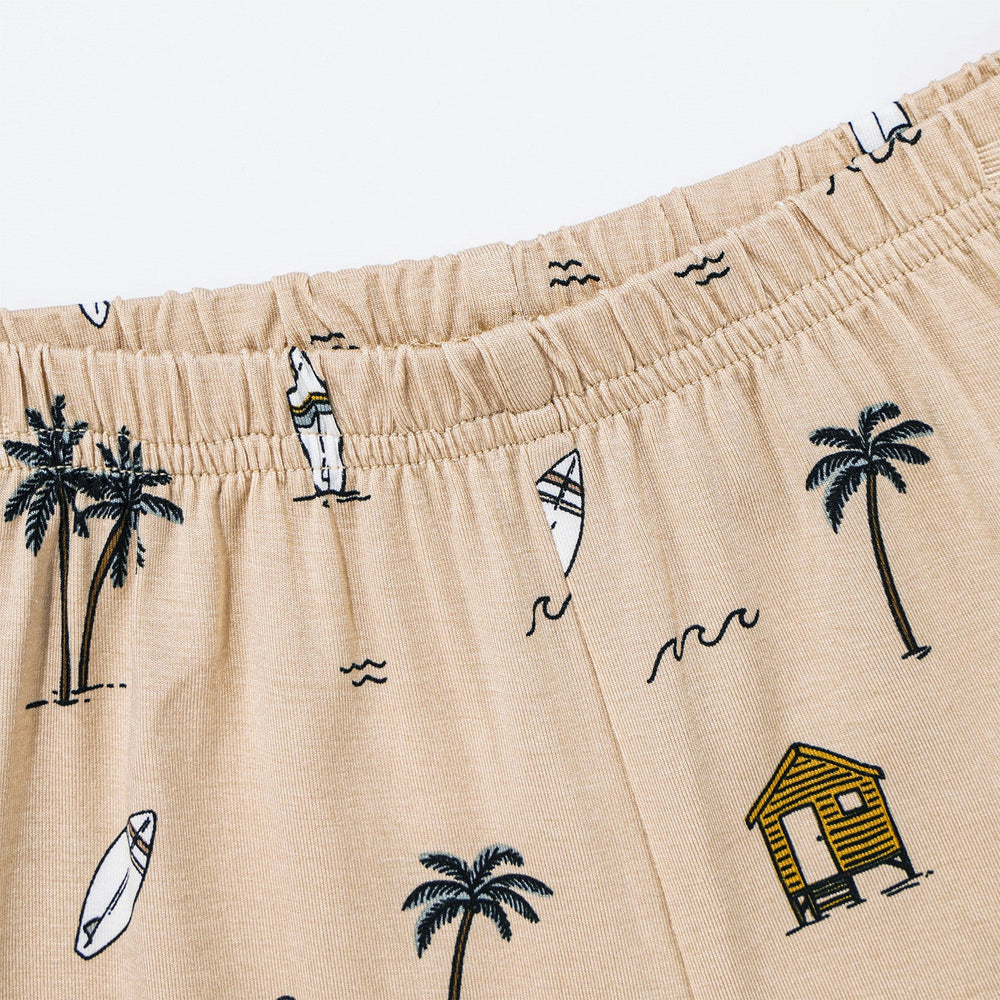 
                  
                    Elastic waistband of tan baby shorts with all-over surf shack and beach motif print – includes palm trees, waves, and surfboards.
                  
                