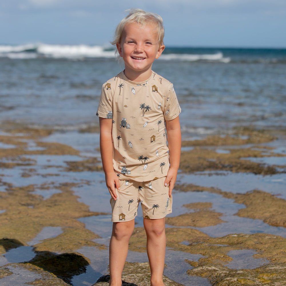 
                  
                    Smiling toddler boy wearing tan Surf Shack two-piece summer outfit with beach illustrations, standing on rocky shoreline.
                  
                
