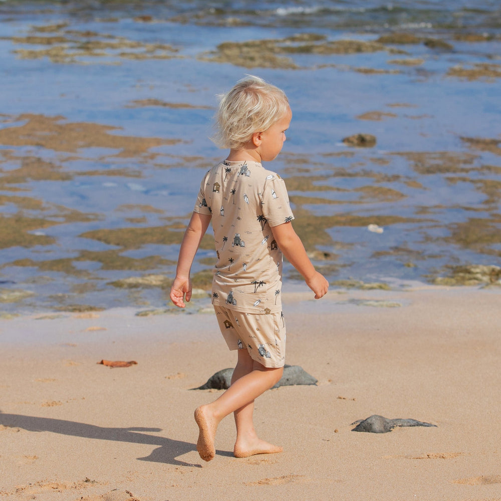 
                  
                    Blonde toddler walking on beach in tan two-piece Surf Shack outfit printed with surfboards, huts, and palm trees – ocean in background.
                  
                