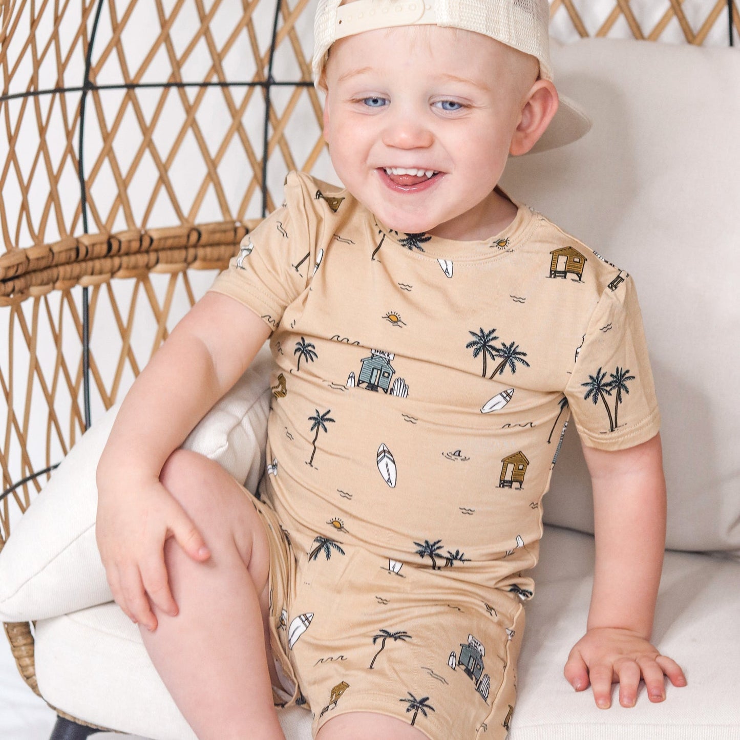 
                  
                    Smiling toddler boy sitting in a rattan chair wearing tan Surf Shack two-piece summer outfit with palm tree and surfboard print.
                  
                